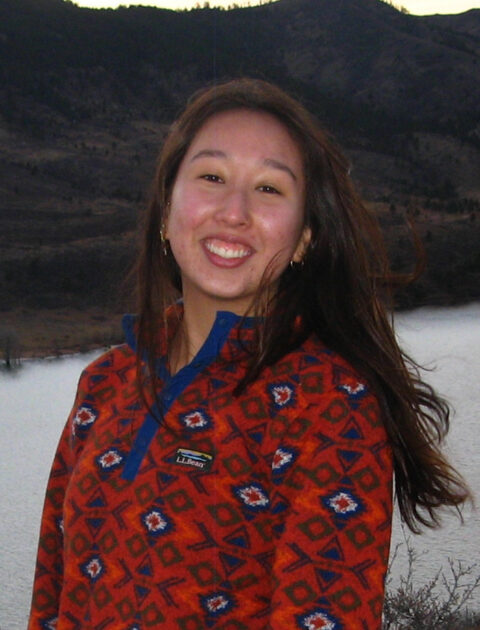 Person smiling outdoors in front of a river and mountains, wearing a red patterned pullover.