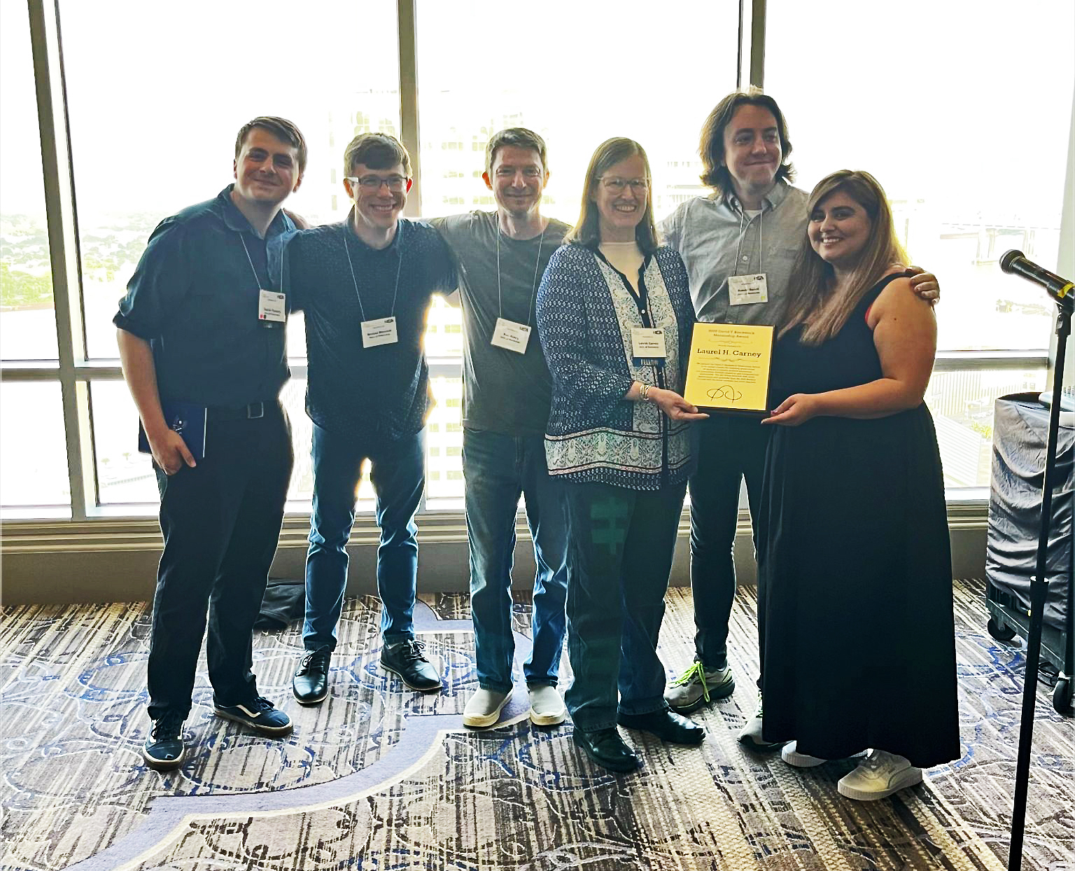 A group of six people smile while presenting an award plaque to University of Rochester faculty member Laurel H. Carney at a conference event.