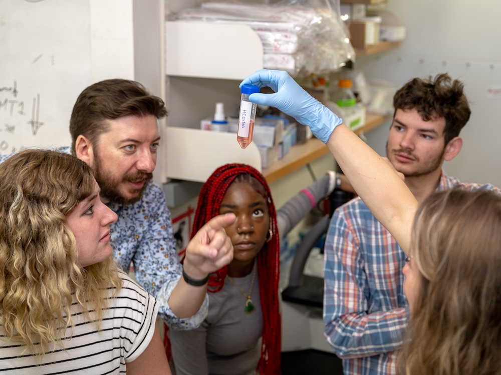 A researcher and students observe a gloved hand holding a test tube in a lab.