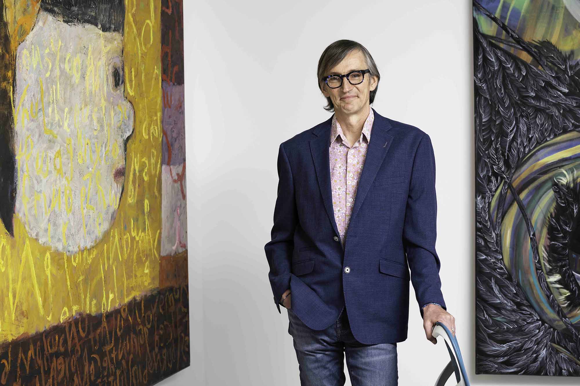 Stephen Dewhurst stands with his hand on a chair between two large paintings.