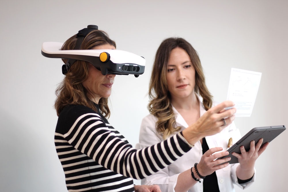 A woman wears an elaborate headset and peers at a sheet of paper while a doctor in a white coat stands next to her with a clipboard.