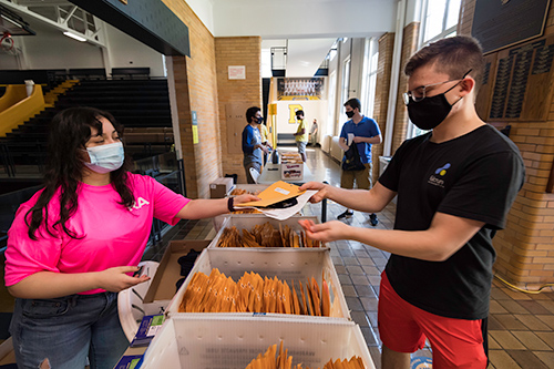 student in a bright RA t-shirt wears a mask and hands an envelope to another student, also wearing a mask.
