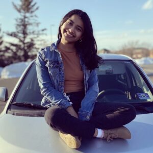 Neha sitting cross-legged on the hood of a car smiling.