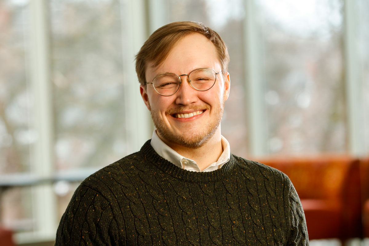 A smiling man with glasses and a sweater poses for the camera, identified as University of Rochester Alumni Bryce Davis ’20.