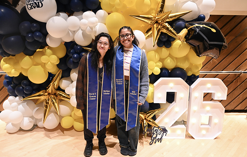 Two students in regalia pose in front of balloons.