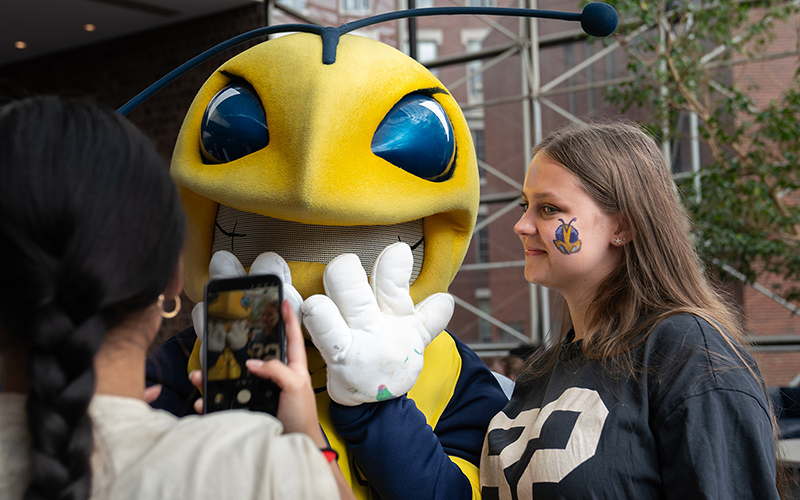 A student with a temporary Rocky face tattoo poses with Rocky for another student taking a photo with their phone