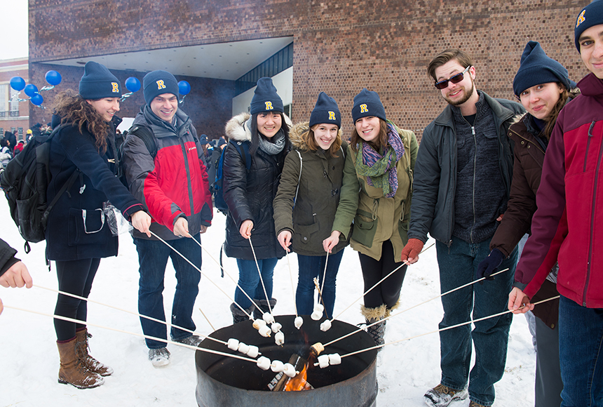 Photo of students posing with marshmallows on sticks around a fire pit on snow-covered Wilson Quad