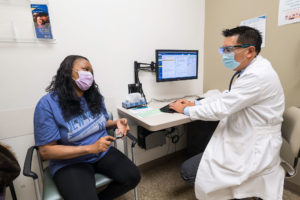 a woman in a blue shirt is sitting in a doctor's office, as the doctor is talking to her while he is on his computer, typing as he talks to her