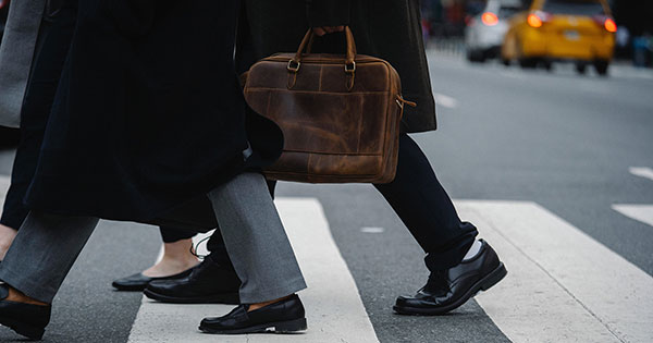 Two men and one woman crossing a New York City crosswalk. Their tailcoats are showing along with their shoes and pants. A brown leather briefcase can be shown held by one of the men. A yellow taxi can be seen in the background, along with a white car ahead of it.