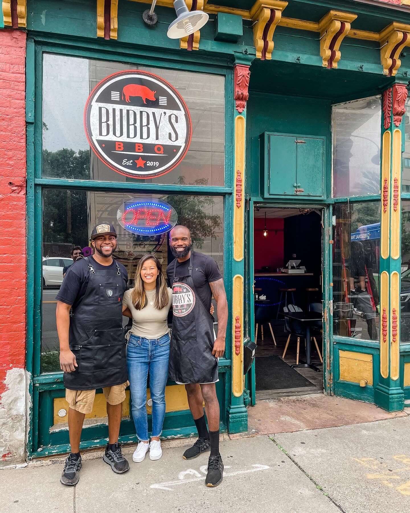 Linh Phillips ’13S (MBA) standing in the middle of two african american men whom work or are the owners of Bubby's BBQ, which they are standing in front of smiling