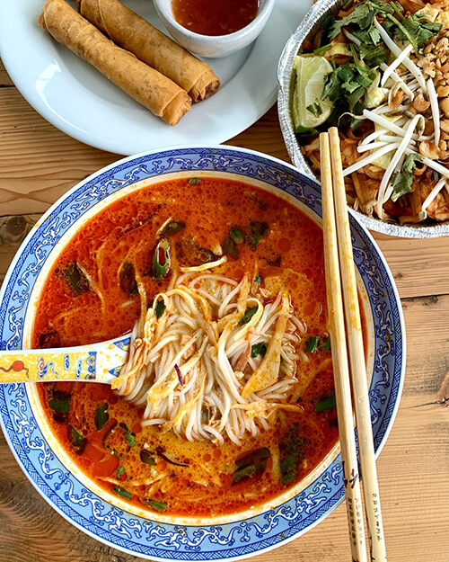 Three dishes of food on a brown wooden table. A set of Chopsticks lay on the main bowl which has a decorative spoon with pasta within it