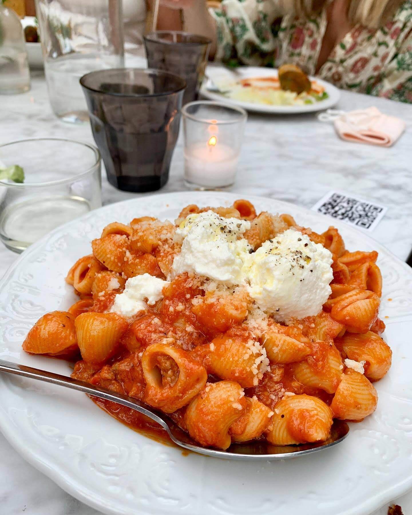 A pasta meal on a white dish, on a table which has a white table cloth, with a woman in the background eating her meal