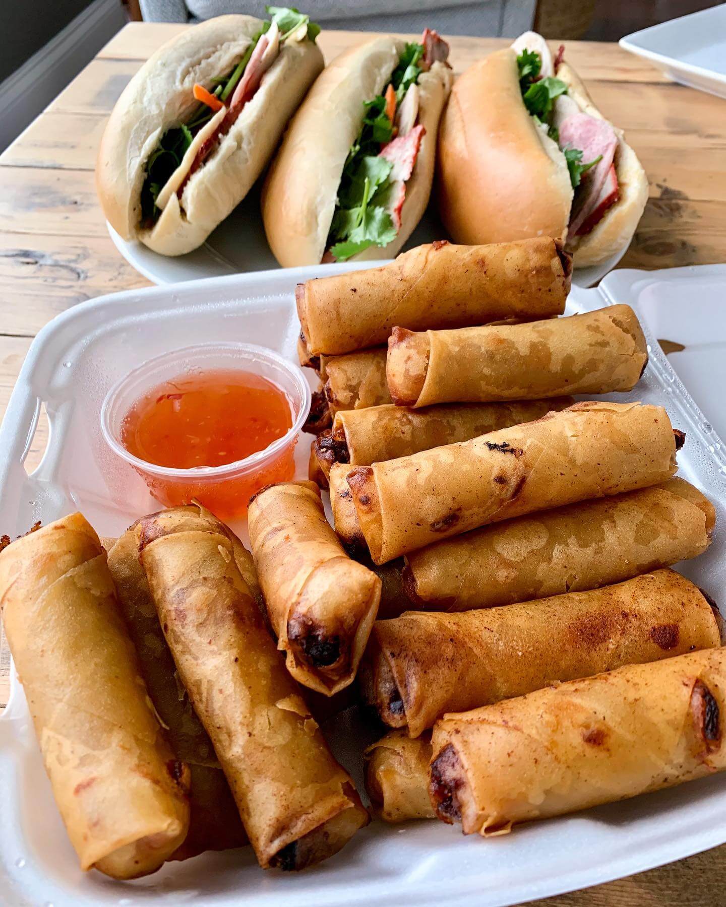 Three sandwiches on the upper section of a table, and twelve egg rolls within a dish next to a orange sauce