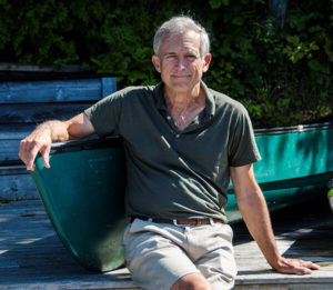 Profile picture of Stephen Plume as he sits down on a outdoor wooden platform wearing a green shirt with beige shorts as he poses with his right arm leaning against a green canoe