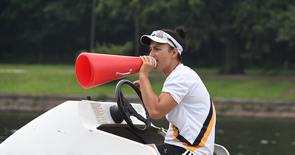 Coach Serra Sevenler using a megaphone in one hand as she steers a boat on a body of water