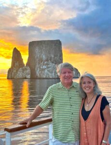 zack and jill stand on a boat with sunset and rock formation seen behind them