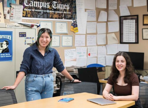 two students pose at a desk in front of The Campus Times poster and newspapers