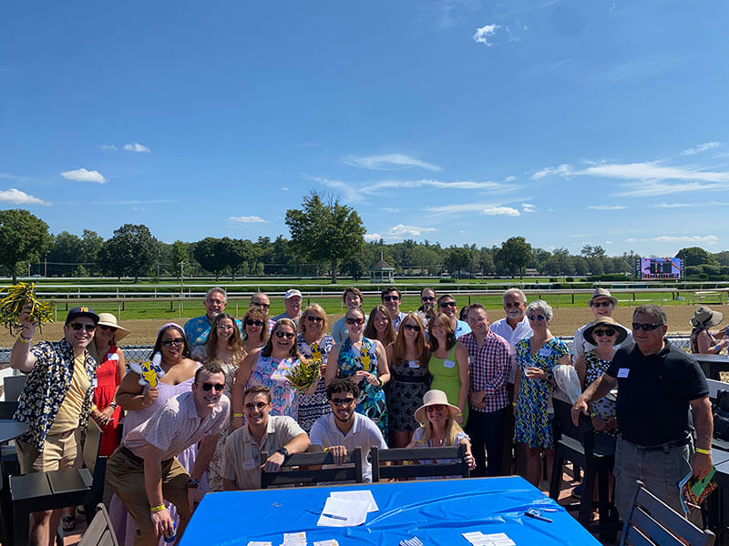 A large group of people pose together in sunny weather at an outdoor event near a horse racing track, smiling and dressed in summer attire.
