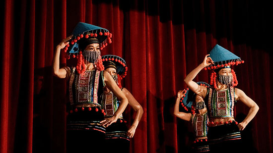 Dancers in vibrant traditional attire perform on stage in front of a red curtain during an Asian Pacific American Heritage celebration.