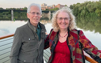George Scharr and his wife, Linda Rice stand on a boat deck by the water with trees and a bridge in the background.