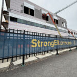A construction worker operates a lift to install exterior panels on a multi-story building under construction behind a fence displaying the StrongER.urmc.edu banner.