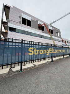 A construction worker operates a lift to install exterior panels on a multi-story building under construction behind a fence displaying the StrongER.urmc.edu banner.