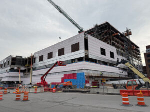Construction crews work on a large multi-story building wrapped in weatherproofing material, with cranes and lifts positioned around the active job site.