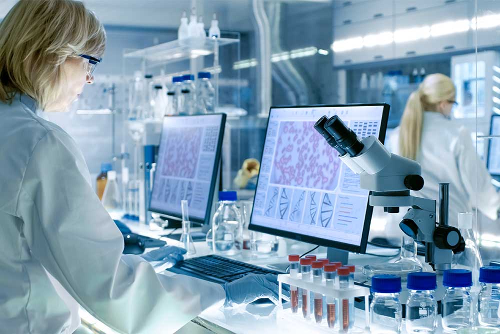two women working in a lab wearing lab coats and protective glasses on computers