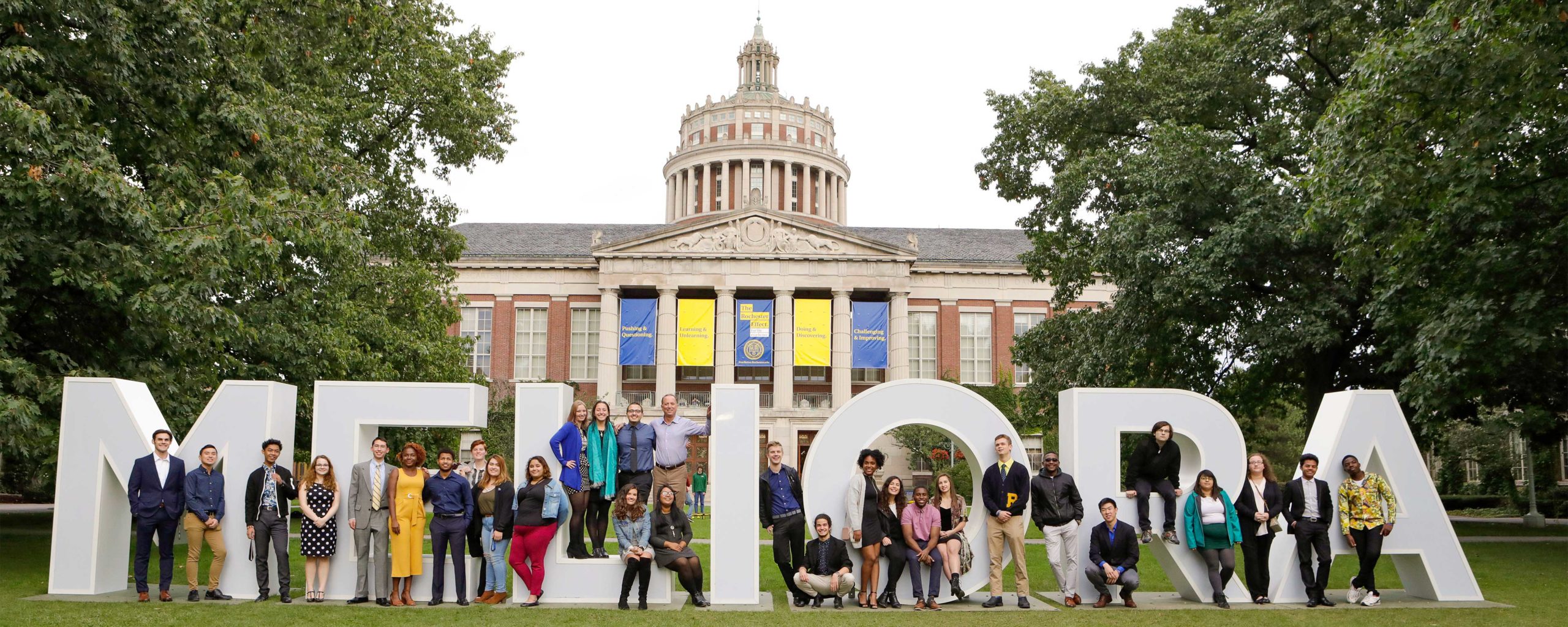 group of handler scholars students standing in front of large meliora letters in quad with rush rhees in background