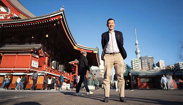 Yohay Wakabayashi standing in front of Tokyo street wtih buildings and people behind him.