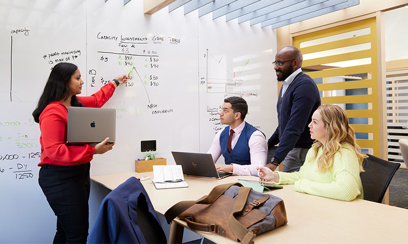 A meeting of 4 people with a woman pointing at a whiteboard, as others watch at conference desk