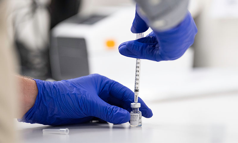 Hands wearing blue gloves pushing a syringe/needle into a vile.