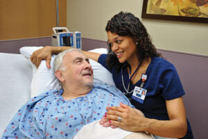 A senior male patient with gray hair is lying in a hospital bed, wearing a hospital gown, and smiling warmly at a nurse beside him. The nurse, a woman with curly dark hair wearing navy blue scrubs and a stethoscope, is holding his hand and engaging in a caring conversation.