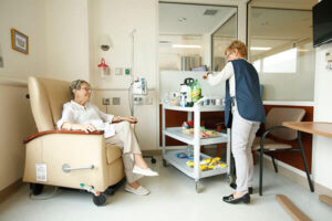 An elderly woman sits in a reclining chair in a hospital infusion center, receiving medical care. A healthcare worker in a blue vest stands nearby, organizing supplies on a mobile cart filled with snacks, drinks, and other essentials.