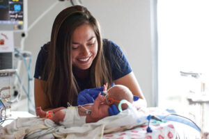 A smiling woman wearing blue medical gloves gently holds the hand of a premature baby in a neonatal intensive care unit (NICU). The baby is lying on a hospital bed, connected to medical tubes and monitors, while the woman provides care and comfort.