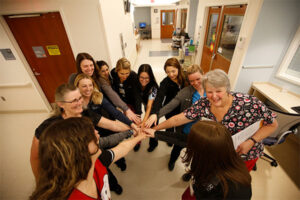 A group of healthcare professionals, mostly women, stand together in a hospital hallway, extending their hands into the center for a team huddle. They are smiling and appear to be celebrating teamwork and unity, with one older woman in floral scrubs holding a clipboard and leading the group.