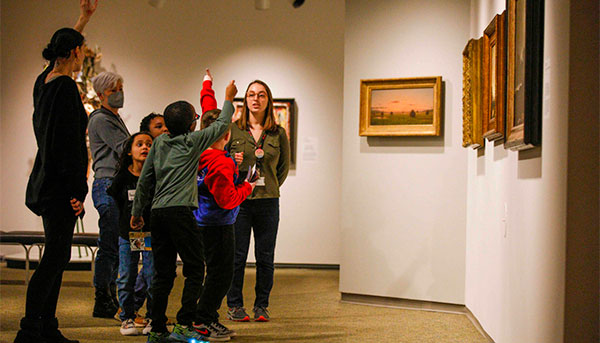 A group of schoolchildren with their hands raised and their Expanded Learrning Collaboration instructors at the Memorial Art Gallery.