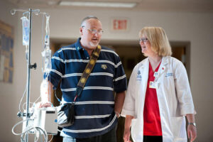 A male patient with glasses, wearing a striped polo shirt, walks through a hospital corridor with an IV drip while talking to a smiling female healthcare professional in a white coat. She has blonde hair, glasses, and an identification badge, and they appear to be engaged in a friendly conversation.