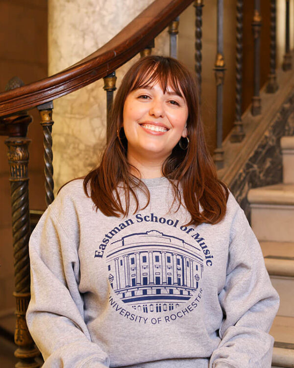 A smiling woman sits on a staircase wearing a gray Eastman School of Music University of Rochester sweatshirt.