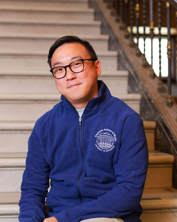 A man wearing glasses sits on a staircase in a blue Eastman School of Music University of Rochester fleece jacket.