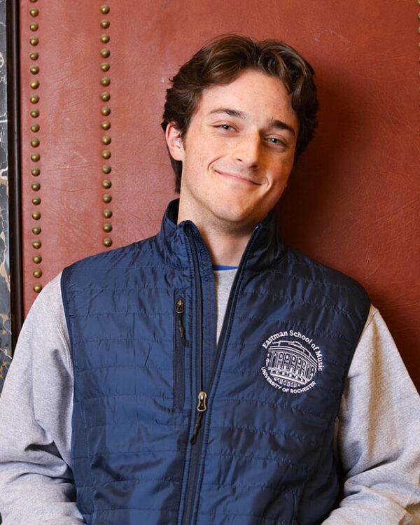 A young man leans against a wall wearing a blue Eastman School of Music University of Rochester vest over a gray sweatshirt.