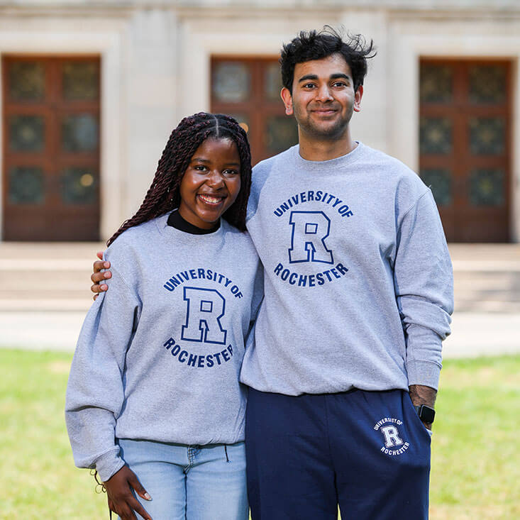 A smiling man and woman wearing matching gray University of Rochester sweatshirts pose together outside on campus.