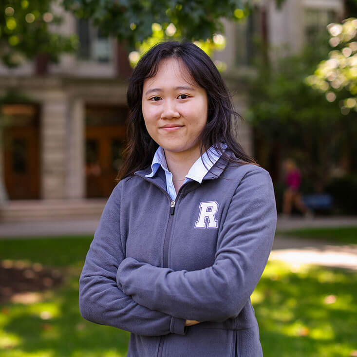 A student stands outdoors with arms crossed, wearing a gray University of Rochester zip-up jacket.
