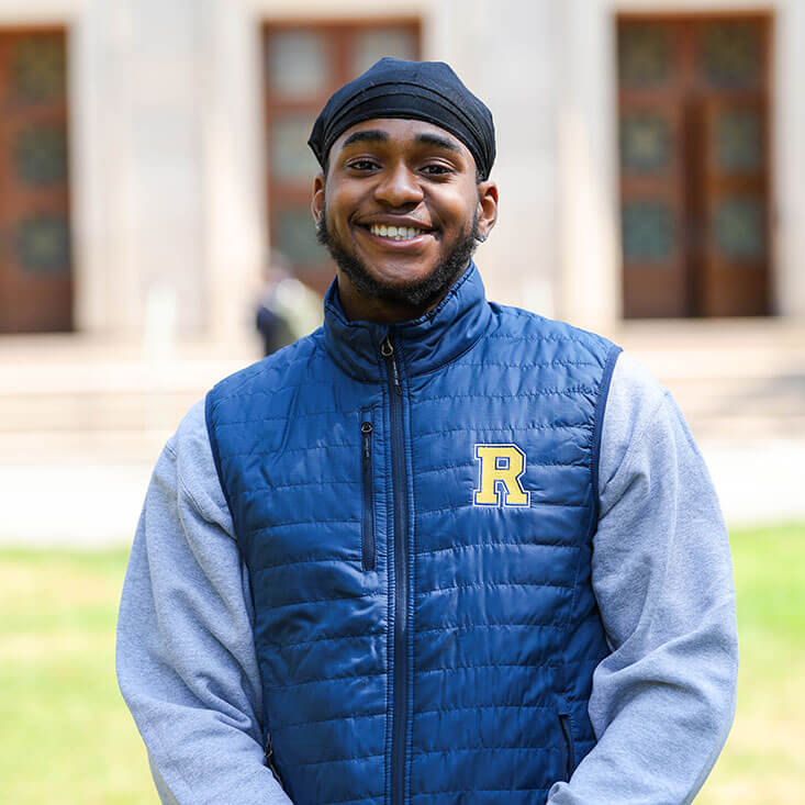 A smiling student wears a blue quilted vest with the University of Rochester “R” logo over a gray sweatshirt.