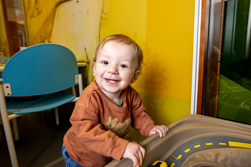 Luke_028_800w A smiling toddler in a rust-colored long-sleeve shirt leans against a play table in a colorful indoor play area with a bright yellow wall.
