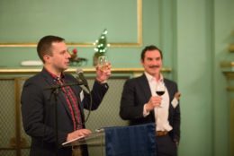 man at podium raising a glass for a toast with another man in background