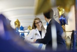 woman wearing glasses wearing a white coat speaking to a man in a suit