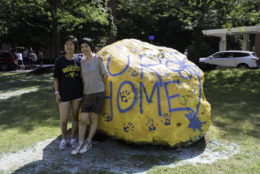 large rock painted yellow with blue UR HOME written on it with two women posing for photo