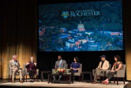 six people on stage with large screen of campus in background