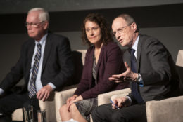 man speaking with hand out wearing a suit next to a woman and a man